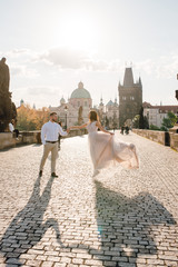 Obraz premium Happy wedding couple of groom and bride in gorgeous dress dancing and smiling while walking on the Charles bridge i the evening on sunset. Prague, Czech Republic