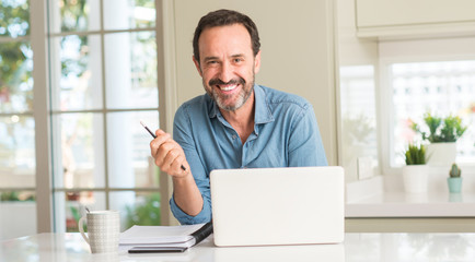Middle age man using laptop at home with a happy face standing and smiling with a confident smile...