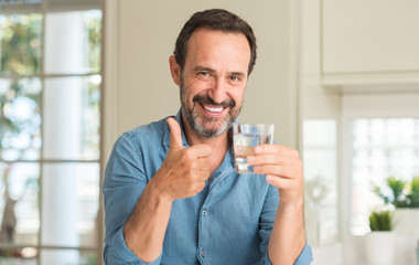 Middle age man drinking a glass of water happy with big smile doing ok sign, thumb up with fingers,...