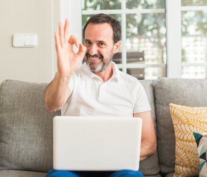 Middle age man using laptop at sofa doing ok sign with fingers, excellent symbol