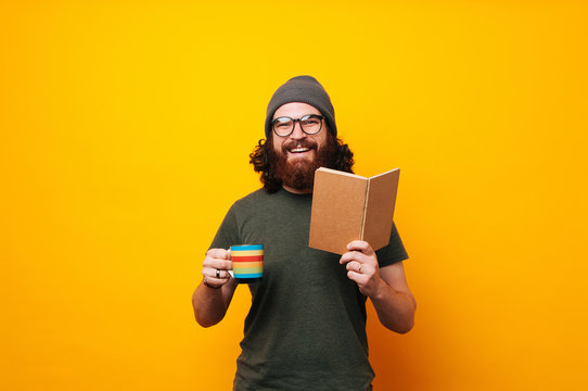 Portrait of a smiling bearded man in eyeglasses reading book and drinking cup of coffee