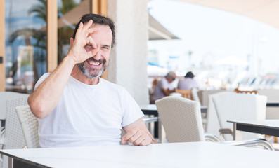 Handsome senior man sitting at terrace with happy face smiling doing ok sign with hand on eye looking through fingers