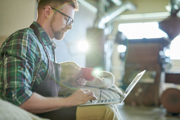 Side view portrait of modern entrepreneur using laptop while sitting on burlap coffee bags in small...