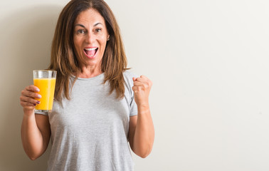 Middle age woman drinking orange juice in a glass screaming proud and celebrating victory and...