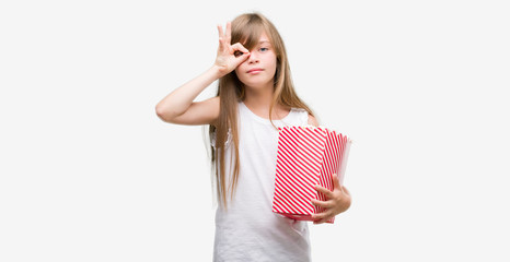 Young blonde toddler holding popcorn pack with happy face smiling doing ok sign with hand on eye looking through fingers