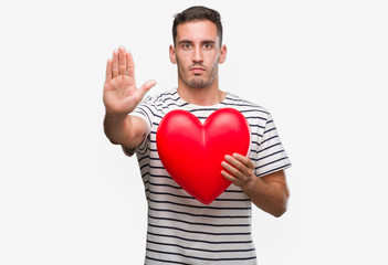 Handsome young man in love holding red heart with open hand doing stop sign with serious and confident expression, defense gesture