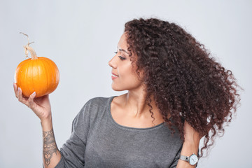 Smiling curly woman holding ripe pumpkin looking at it, touching her hair playful, isolated over...
