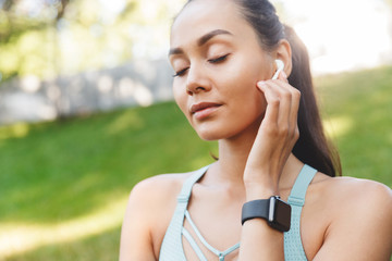 Image closeup of pretty satisfied woman 20s in sportswear using bluetooth earphone and listening to music with closed eyes, during walk in green park