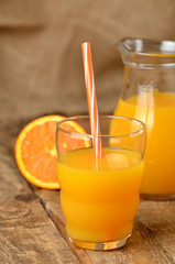 Glass with orange juice and straw, jug with fresh juice and pile of oranges in the background on wooden table - vertical photo