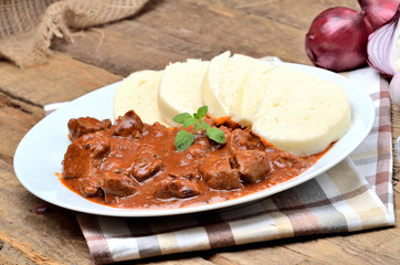 Pork goulash meat with dumplings on white plate, cutlery, garlic, onion, pepper, tablecloth in the background - typical Czech food