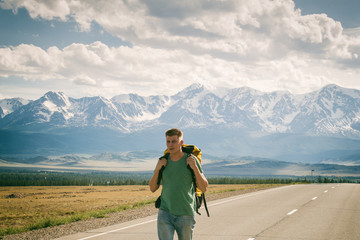 Man walks along an asphalt road with a backpack on his back. Mountain background