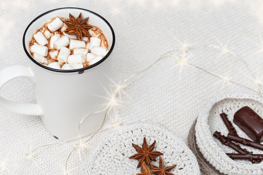 White cup of hot chocolate with marshmallows and sweets and decorated glowing garland on cozy knitted background. Christmas concept.  Selective focus. Copy space.