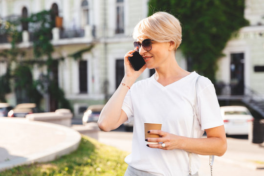 Image of caucasian blond woman wearing white t-shirt and sunglasses walking through city street in summer with takeaway coffee, and speaking on mobile phone
