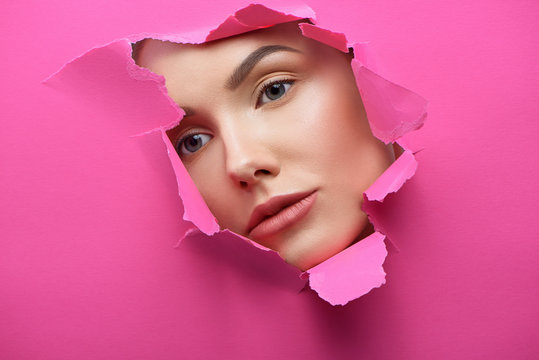 Pretty girl's face rupturing pink cardboard. Stiff, thick carton making contrast with model face, having plump lips, big eyes,healthy, smooth skincolor. Looking at side. Looking bohemic and beatiful.