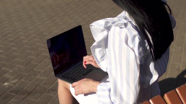Brunete typing on laptop sitting on a bench in park