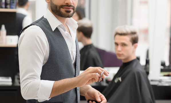 Cropped photo of hairdresser working in barbershop using scissors. Wearing white classic shirt, grey waistcoat. Making fashionable, stylish haircut for young boy reflected in mirror on background.