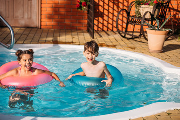 portrait of little siblings with inflatable rings swimming in swimming pool together on summer day