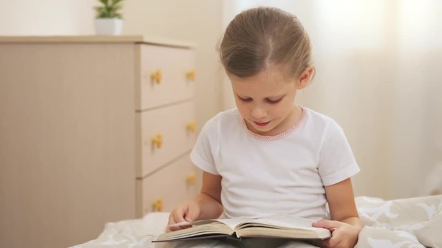 Cute little girl sitting on bed with textbook sunny day. Child learning to read. 