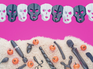 Sex toys are on sheepskin fur. Next to them are scattered artificial pumpkins. From above there is a black and white garland in the form of skulls. There is a fuchsia-colored background.