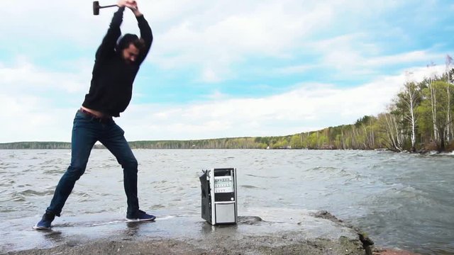 Concept of freedom from computer addiction - Man breaks system unit of a computer with a hummer outside, lake with forest background. Angry man destroying a laptop or computer with a hammer.