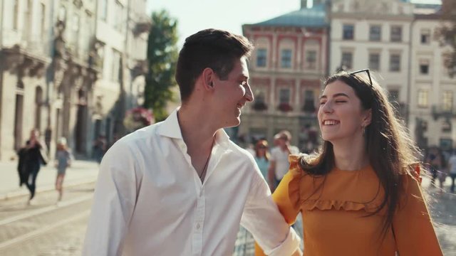 Sweet young people in love walk down the historical city center, holding their hands, laughing, a man kisses his girlfriend in cheek. Couple goals, true feeling, joy of life. Love story