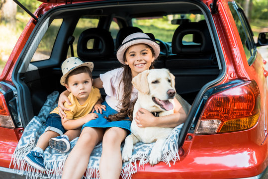 siblings sitting on car trunk with labrador dog