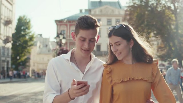 Handsome young man embracing waist of his cheerful attractive young girlfriend actively uses his smartphone, while they&rsquo;re walking down the central square. Romanticism, social networks. Love story