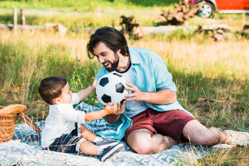 handsome father and toddler son playing with football ball at picnic
