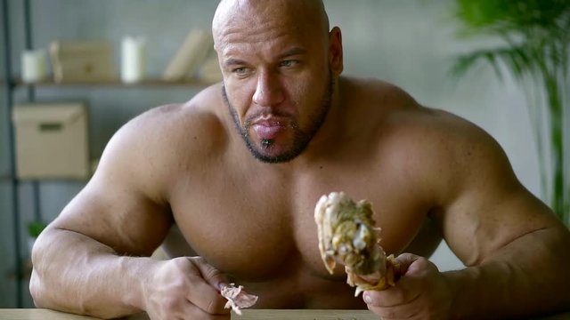 Portrait of a muscular, brutal, bald naked man biting and chewing a big cooked turkey leg.