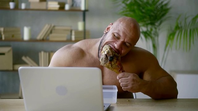Front view shot of shirtless muscular man sitting at the table behind computer and eating big, fat turkey leg.