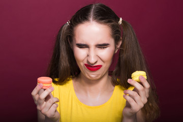 Upset dieting brunette girl with bright makeup holding macaroons. Studio shot on a red background