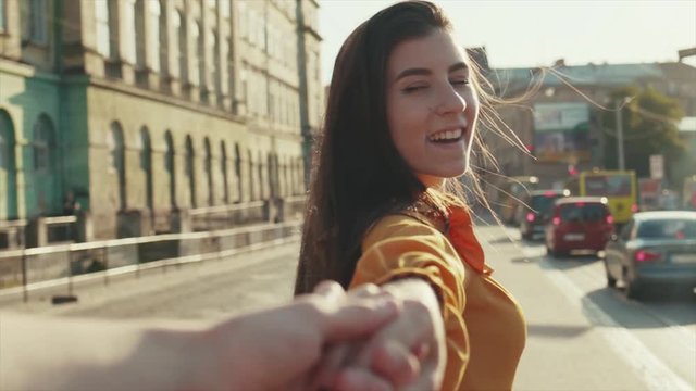 Attractive long-haired woman holds male hand, leads his forward to the city center. Dating, couple goals, happy together. Windy weather, sunlight. Love story