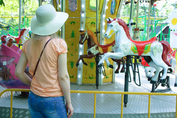 A girl in a straw hat in an amusement park looks at the merry-go-round.