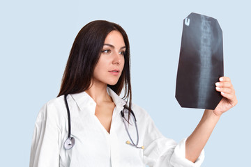 Serious female doctor looks attentively at X ray, examines people`s spine, wears white lab gown,...