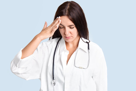 Attractive brunette young female nurse has headache after making diagnosis, keeps hand on forehead, concentrated down, dressed in white robe, isolated over blue background. Overworked doctor