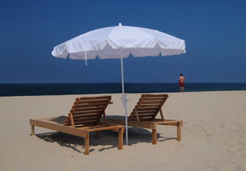 Espreguiçadeiras na praia - com um guarda-sol branco e uma pessoa do sexo masculino ao longe com calções vermelhos