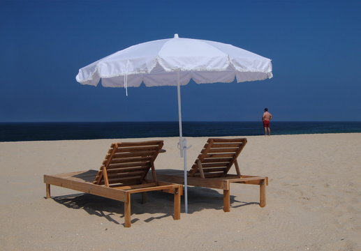 Espregui&ccedil;adeiras na praia, com um guarda-sol branco e uma pessoa do sexo masculino ao longe com cal&ccedil;&otilde;es vermelhos