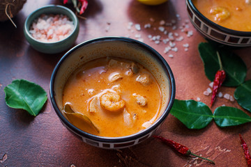 Tasty asian soup tom yam in small bowls, chilli pepper, salt and coconut and lime on a dark brown background. Healthy organic food concept, selective focus