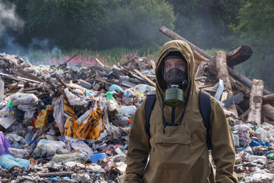 A working man in a gas mask against the backdrop of burning garbage. A lot of plastic bags thrown to the dump. From the plastic debris goes smoke. On the man, the khaki color encephalic suit