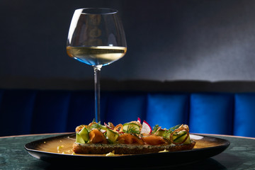 Smorrebrod Danish sandwich with salmon fish, curd mousse, marinated cucumber and radish. Delicious vegetarian snack food with dark rye bread and different topping and wine glass at restaurant table