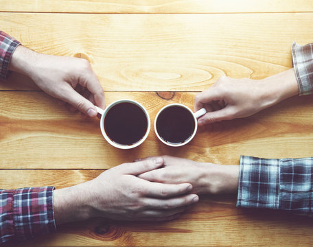 Hands of couple with two cups of morning coffee on wooden table. View from above
