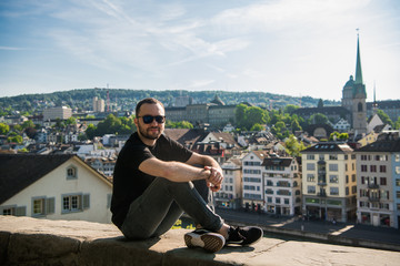 A handsome man wears a nice sunglasses and looking to the city. An attractive mid-aged guy is sitting on a enbankment of a beutiful european city. A cool guy feels so relaxed and happy when see some