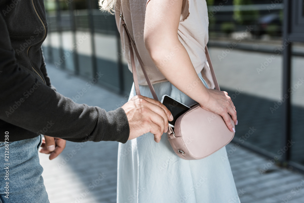 Wall mural cropped view of criminal man pickpocketing smartphone from womans bag on street