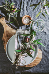 Top view of table setting with gray Linen tablecloth and napkin, white plate, cutlery and and olive tree branch boards decoration. Top view