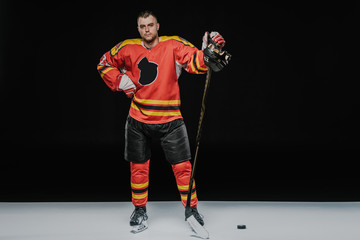 Fototapeta premium full length view of professional ice hockey player standing with hand on waist and looking at camera on black