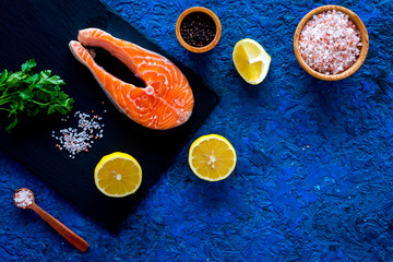 Preparing salmon steak with spices, seasoning. Piece of fresh fish on cutting board near sea salt, lemon slices, greenery on blue background top view copy space