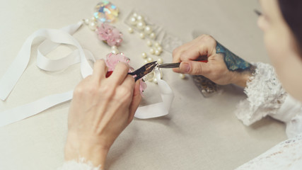 Girl fashion designer makes jewelry. Shooting close-up. She makes a hook on a pin with artificial pearls. Ribbons and artificial flowers are on the table