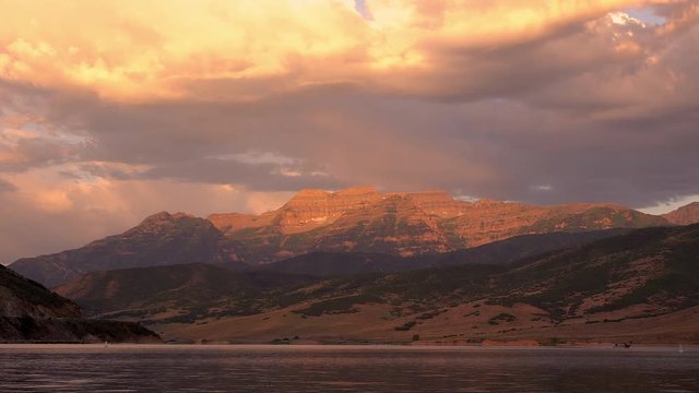 Timpanogos Mountain lit up at sunrise with colorful clouds above as boat moves across Deer Creek Reservoir.