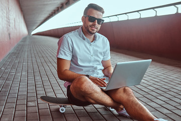Portrait of a happy handsome fashionable freelancer in sunglasses dressed in a white shirt and shorts working on a laptop while sitting on a skateboard under a bridge, looking at a camera. © Fxquadro