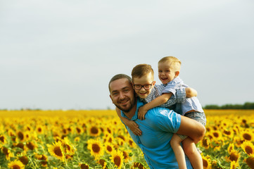 Fototapeta premium Cheerful father with his sons on vacation in the field with sunflowers . The parent plays with his son . Father's day